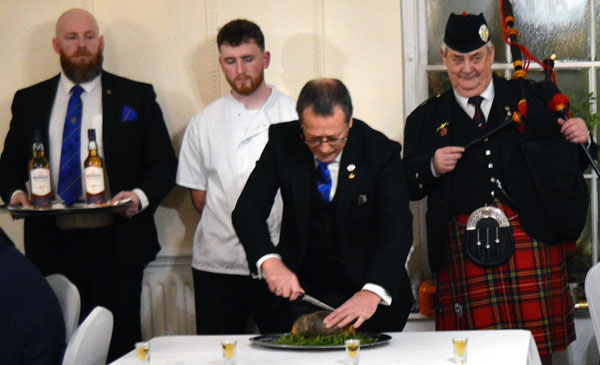 Gordon Maclellan stabbing the Haggis during his address, accompanied by a lodge steward, the chef, and the piper.